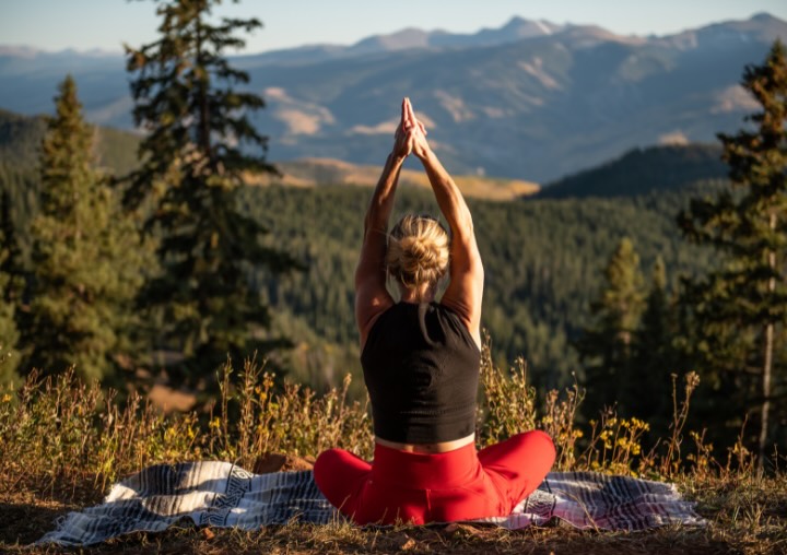 Person practicing yoga outdoors with hands raised, surrounded by mountains and trees, emphasizing a mind-body connection for holistic health.