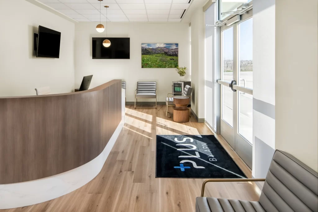 Reception area of APN Dallas-Fort Worth, featuring a curved wooden front desk, modern seating, and a welcoming atmosphere, with the APN Plus logo on the floor mat, emphasizing care and support for Intensive Outpatient Programs.