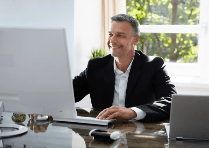 Smiling man in a suit working at a desk with a computer, reflecting a professional environment at All Points North Dallas-Fort Worth addiction treatment center.