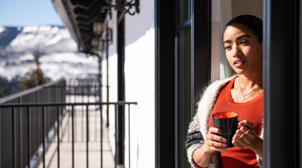 Woman holding a cup, looking thoughtfully from a balcony with a snowy mountain backdrop, symbolizing resilience and reflection in a tranquil setting.