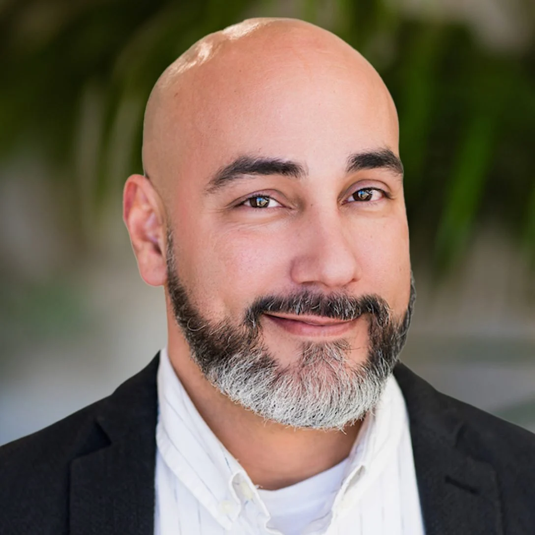 Bald man with a beard smiling, wearing a black blazer and white shirt, representing the APN DFW staff in a professional setting.